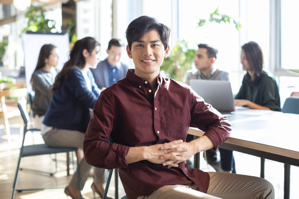A young business intern looking confident while his team is meeting in the background