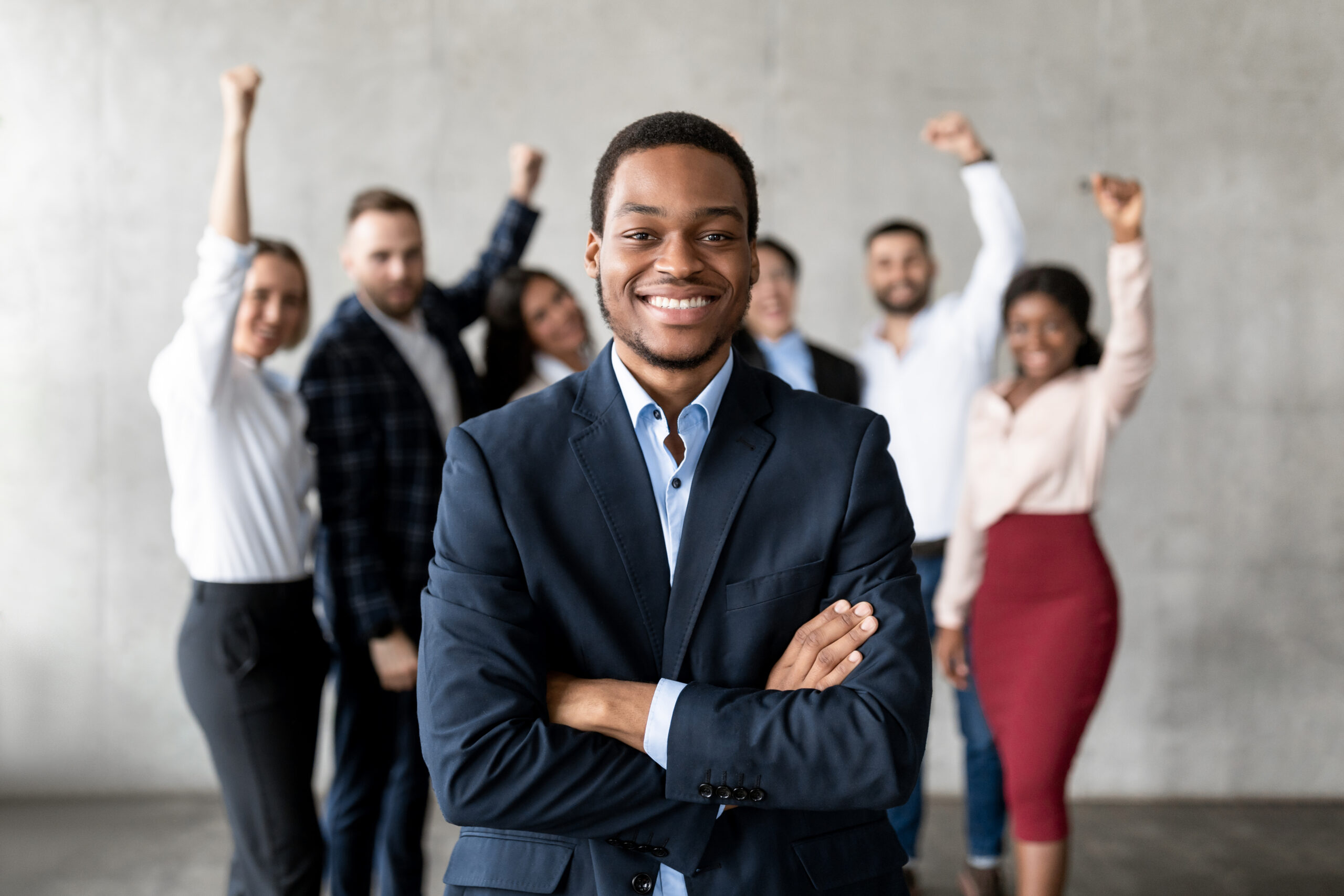 A smiling young professional with his arms crossed and his team in the background, raising their hands to cheer for him.